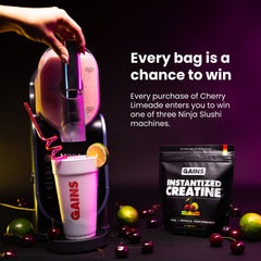 A hand inserts a capsule into a black and pink Gains Instantized Creatine machine, which dispenses a drink into a white Gains cup. A black bag of Gains Instantized Creatine is shown next to the machine on a dark background with cherries and limes scattered around | cherry limeade