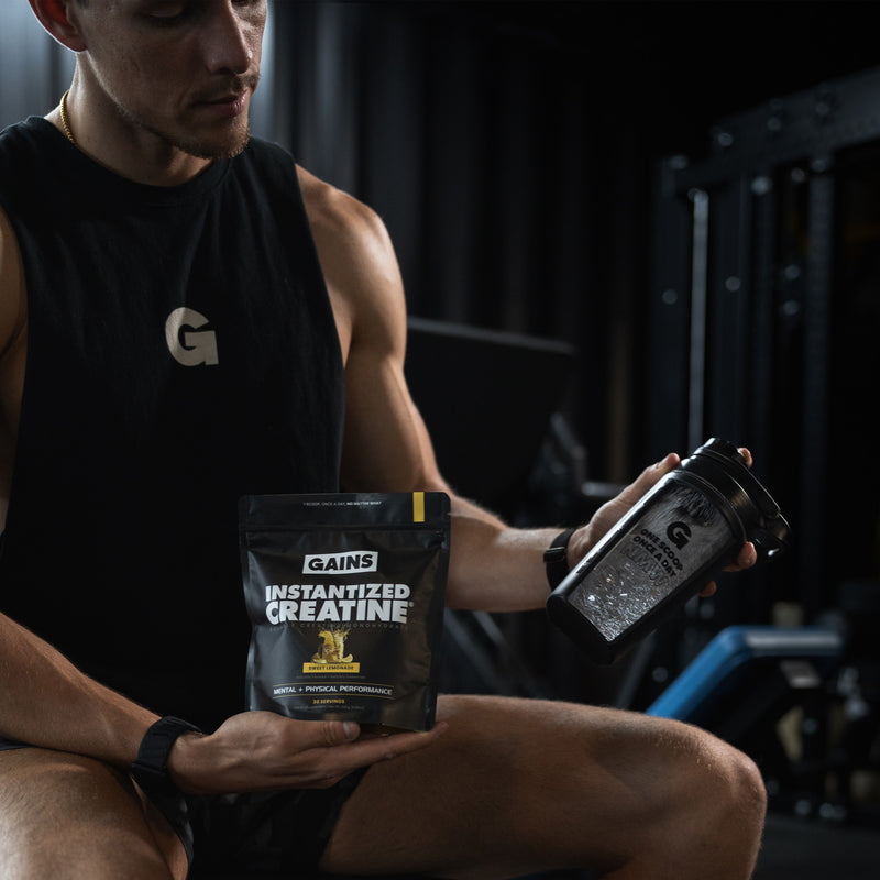 Man holding a package of Gains Instantized Creatine and a shaker bottle in a gym setting. | sweet lemonade 30