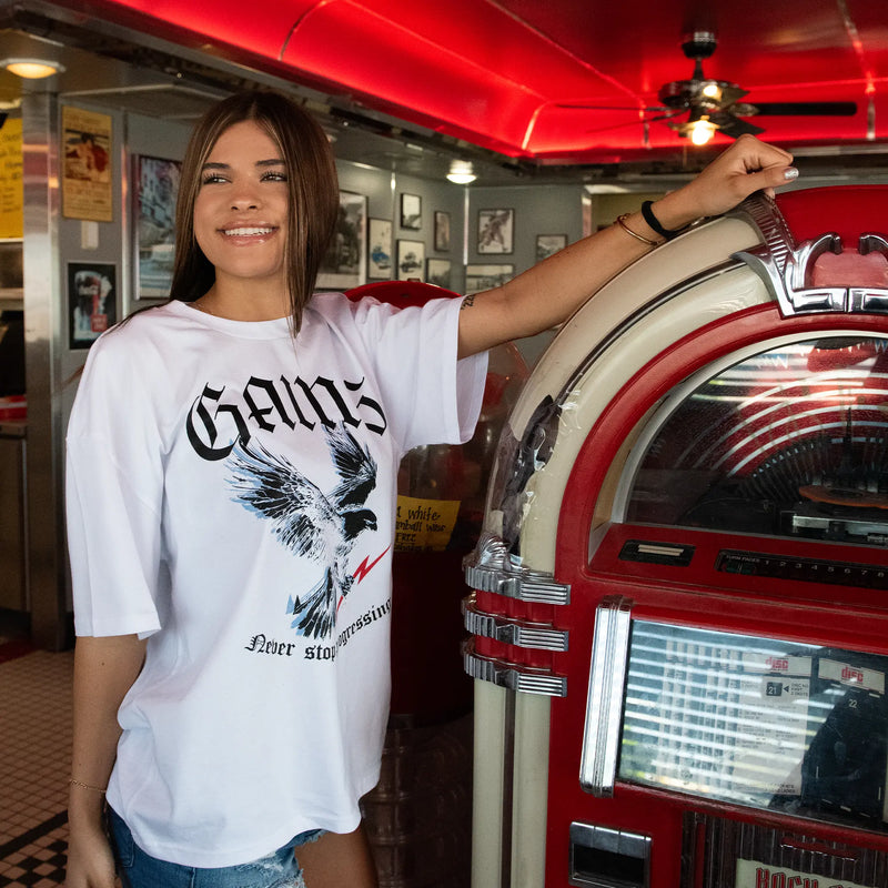 Woman in a white t-shirt with an eagle design standing next to a red and white jukebox in a retro setting. Gains USA 250th Anniversary T-shirt 