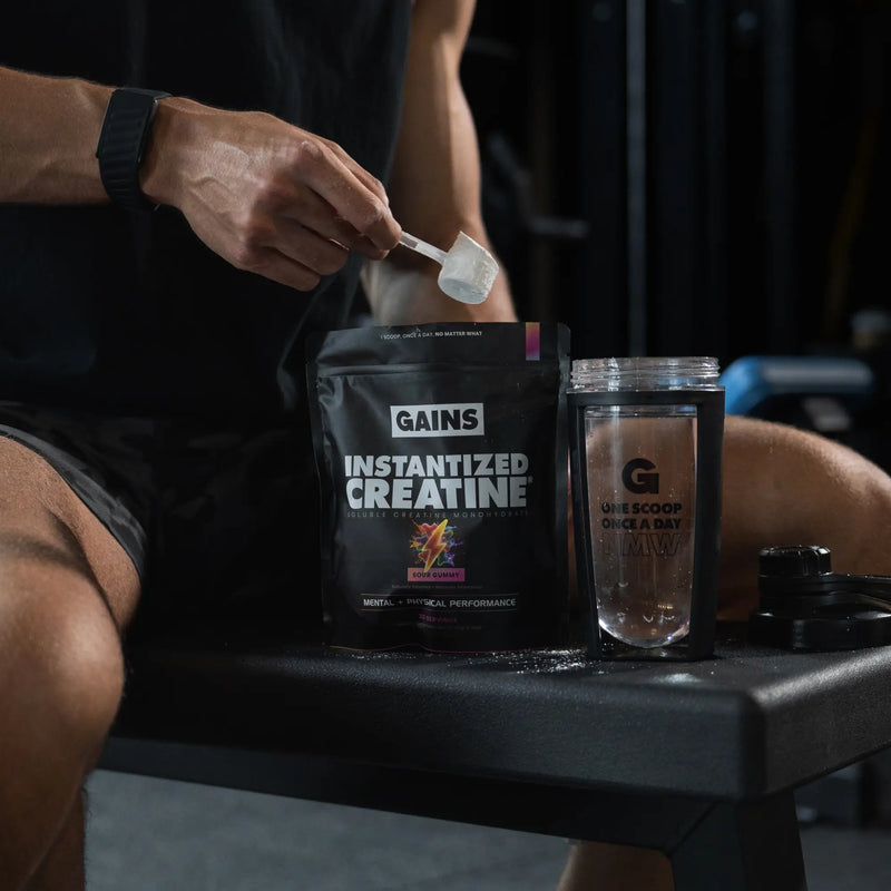 Person holding a scoop of Gains Instantized Creatine next to a glass container with a supplement, sitting on a bench. | sour gummy