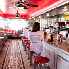 Person sitting at a bar counter in a diner with red seats and a red ceiling. Gains USA 250th Anniversary T-shirt 