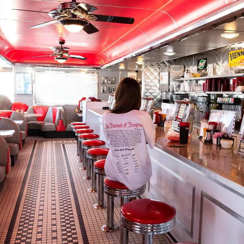 Person sitting at a bar counter in a diner with red seats and a red ceiling. Gains USA 250th Anniversary T-shirt 