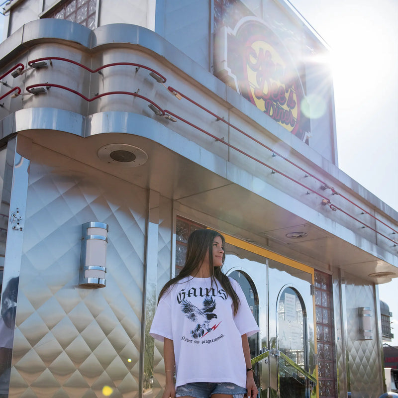 Woman wearing Gains in Bulk USA t-shirt standing in front of 50s diner. 